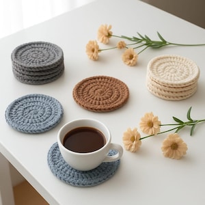 Può includere: Un set di sottobicchieri all'uncinetto fatti a mano in vari colori, tra cui grigio, marrone e blu. Una tazza bianca di caffè è appoggiata su un sottobicchiere blu. I sottobicchieri sono disposti su una superficie bianca con fiori decorativi.