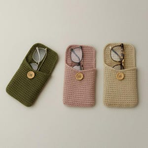 May include: Three crochet eyeglass cases in olive green, dusty rose, and beige. Each case has a wooden button closure and holds a pair of eyeglasses. The cases are handmade and have a textured appearance.