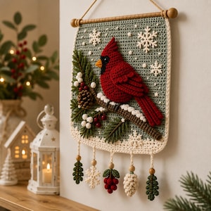 Op de afbeelding: Een gehaakte wandhanger met een rode kardinaalvogel op een bruine tak met groene dennennaalden, rode bessen en een dennenappel tegen een saliegroene achtergrond met witte sneeuwvlokken en vijf bungelende ornamenten aan de onderkant.