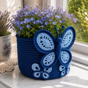 May include: A dark blue crocheted basket featuring a large, light blue and dark blue butterfly appliqué on the front, along with two smaller flower appliqués at the base. The basket is filled with a dense cluster of small, vibrant purple bell-shaped flowers.