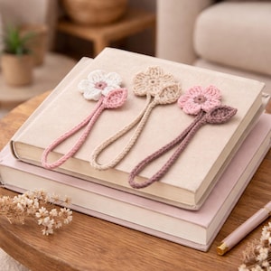 May include: Three handmade crochet flower bookmarks in white, beige, and pink, each with a matching leaf and cord. The bookmarks rest on a stack of books with a neutral colour palette, suggesting a cosy reading theme.