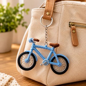Puede incluir: Un llavero de bicicleta tejido a ganchillo con marco azul claro, asiento y manillar marrones, y neumáticos negros con radios plateados. El llavero está unido a una anilla y cadena de metal plateado, colgado de un bolso beige texturizado.