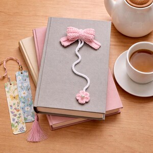 May include: A grey hardback book with a pink crocheted bow bookmark. The bookmark has a white cord and a pink flower. Two floral bookmarks and a pink tassel are also visible. A teapot and cup of tea are in the background.