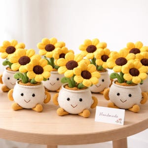May include: A collection of handmade crochet sunflower planters. Each planter features a smiling face, arms, and legs. The sunflowers have yellow petals and brown centers. A small card with the word "Handmade" is visible.