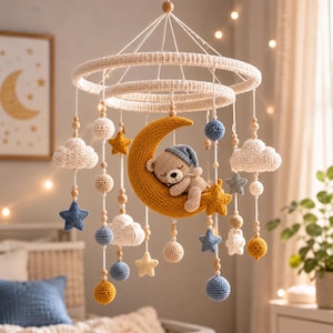 Puede incluir: Un móvil de ganchillo para bebé con una luna creciente amarilla central y un osito de peluche durmiendo con un gorro azul. El móvil incluye nubes blancas, estrellas azules y amarillas, y pequeñas esferas en azul, amarillo y crema, suspendidas de dos anillos circulares blancos.