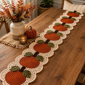Puede incluir: Un camino de mesa de ganchillo largo y rectangular con un patrón repetitivo de calabazas naranjas con tallos marrón oscuro y hojas verdes, bordeado por un borde festoneado de color crema. El camino está hecho de hilo con textura.