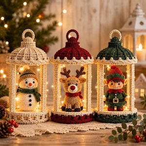Puede incluir: Tres adornos de linterna de ganchillo con un muñeco de nieve, un reno y un elfo. Cada linterna tiene un lazo en la parte superior y contiene una pequeña figura iluminada por luces. Las linternas son de color crema, rojo y verde oscuro.