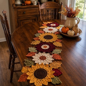 May include: A crochet table runner featuring a series of interconnected floral motifs in shades of yellow, cream, burgundy, and brown, interspersed with crocheted autumn leaves in green, orange, and red, arranged in a linear pattern on a wooden table.