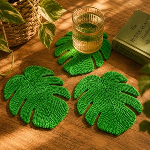 May include: Three bright green crochet coasters shaped like monstera leaves with detailed ribbed texture. One coaster supports a clear glass filled with a light-colored beverage, while the other two lie flat, showcasing the leaf-like design.