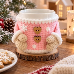 May include: A white ceramic mug featuring a pink crocheted cozy with white snowflake accents, two wooden heart-shaped buttons, and two beige crocheted mittens attached to the sides, creating a festive winter-themed drinkware accessory.