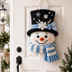 May include: A crocheted snowman wall hanging featuring a black top hat adorned with white snowflakes, a light blue band, and blue berries. The snowman has a white face, black eyes, a carrot-orange nose, pink cheeks, and a blue and white striped scarf.