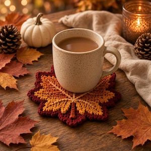 May include: A beige speckled ceramic mug filled with a light brown beverage sits on a crocheted coaster shaped like a maple leaf. The coaster features a gradient of colors including deep burgundy, vibrant orange, and golden yellow yarn.