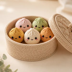 Puede incluir: Una cesta redonda de ganchillo beige con tapa a juego que contiene cinco pequeños peluches esféricos de ganchillo. Cada peluche presenta una cara sonriente con ojos negros, boca cosida y mejillas rosadas, en rosa claro, verde, amarillo, blanco y naranja.
