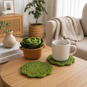 May include: A cozy living room scene features a handmade crochet cabbage in a small basket, a white mug, and two green crochet coasters. The items are arranged on a wooden coffee table, creating a warm and inviting atmosphere.
