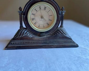Puede incluir: Un reloj de repisa decorativo de madera con esfera redonda y números romanos. El reloj tiene un bisel dorado y un marco y base de madera marrón oscuro. El reloj mide aproximadamente 15 cm de alto.