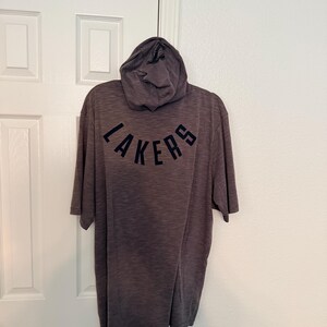 May include: A gray hooded t-shirt with the word "LAKERS" arched across the back in navy blue lettering. The hood has a small logo on it. The shirt is hanging on a white door.