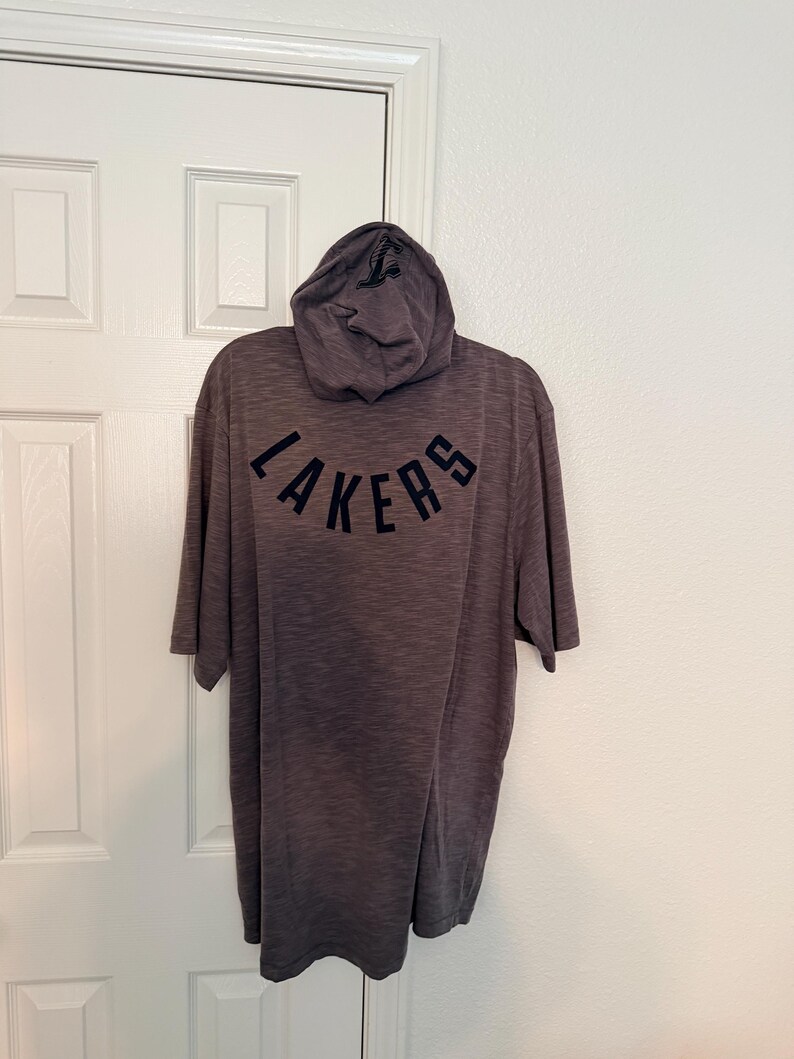May include: A gray hooded t-shirt with the word "LAKERS" arched across the back in navy blue lettering. The hood has a small logo on it. The shirt is hanging on a white door.
