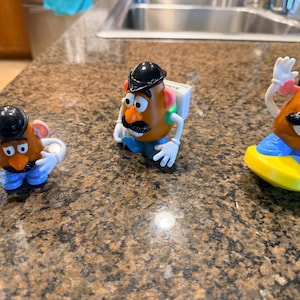 May include: Three Mr. Potato Head toys are arranged on a speckled brown countertop. Each toy has a brown potato-shaped head, a black hat, and a large orange nose. They wear blue pants and have white hands. One is waving, another has a backpack.