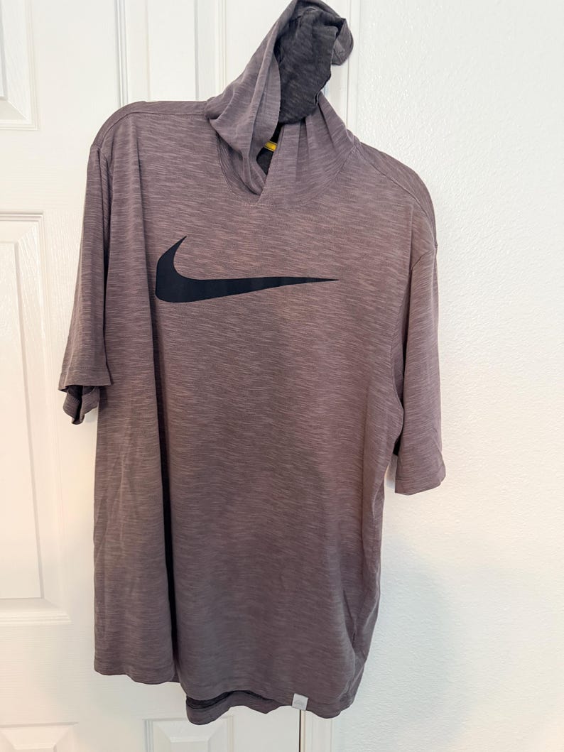 May include: A gray hooded shirt with short sleeves, featuring a black Nike swoosh logo on the front. The shirt has a textured appearance and a contrasting dark gray hood.