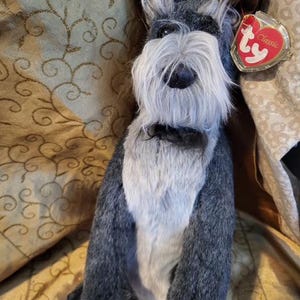 May include: A plush Schnauzer dog with gray and white fur, a long white beard, and black eyes. It has a black collar and a red heart-shaped tag that says "Classic". The dog is sitting on a gold-colored patterned fabric.