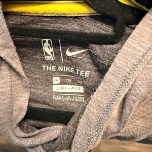 May include: Gray NBA Nike tee shirt with a yellow collar detail. The shirt features the NBA logo and Nike's swoosh. The text "THE NIKE TEE" and "DRI-FIT" are printed on the shirt. The shirt is size XXL.