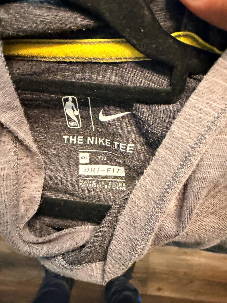 May include: Gray NBA Nike tee shirt with a yellow collar detail. The shirt features the NBA logo and Nike's swoosh. The text "THE NIKE TEE" and "DRI-FIT" are printed on the shirt. The shirt is size XXL.