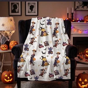 Puede incluir: Una manta blanca con un patrón repetitivo de varios personajes de dibujos animados vestidos con disfraces de Halloween, incluyendo figuras con sombreros de bruja y trajes de vaquero, junto a pequeñas ilustraciones de murciélagos, fantasmas y calabazas.