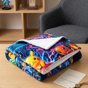 以下が含まれることがあります： ピカチュウとヒトカゲが描かれた鮮やかなグラフィックプリントの折り畳まれたプラッシュブランケット。青い稲妻と紫の回路パターンが特徴です。ブランケットには柔らかい白いシェルパフリースの裏地が付いており、模様が見えるように折り畳まれています。