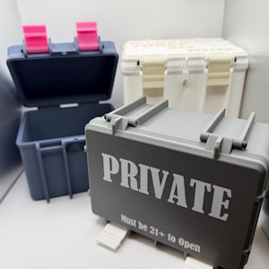 May include: Three small storage containers. One is dark gray with the word "PRIVATE" in white and the text "Must be 21+ to open". Another is navy blue with pink latches, and the third is white.