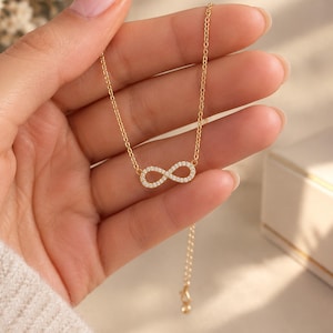 Könnte beinhalten: Eine goldfarbene Halskette mit einer feinen Kette und einem zentralen Anhänger in Form eines Unendlichkeitssymbols. Das Unendlichkeitssymbol ist mit kleinen, klaren, rund geschliffenen Steinen besetzt, die das Licht reflektieren.
