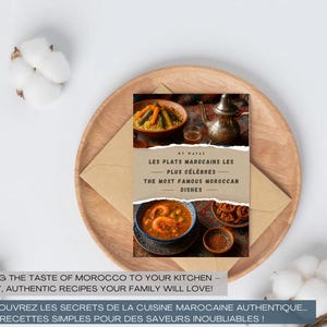 Peut inclure: Un livre de cuisine intitulé "Les Plats Marocains Les Plus Célèbres" est posé sur une assiette en bois, avec une enveloppe en dessous. La couverture présente des plats marocains. Le texte dit "Apportez le goût du Maroc dans votre cuisine".
