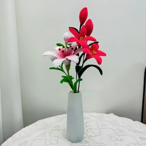 May include: A display of artificial lilies in a white vase. The flowers are a mix of pink and red, with green stems and leaves. The vase is on a white lace tablecloth.