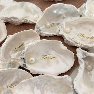 Könnte beinhalten: Eine Sammlung weißer Austernschalen, jede mit einer Perle und einem Namen in goldener Schrift. Die Schalen sind auf einer strukturierten Oberfläche angeordnet. Die Namen lauten "Lora", "Xenia", "Tassos" und "Eva".