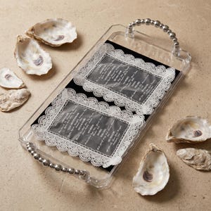 May include: A rectangular clear acrylic serving tray with silver-toned beaded handles. Inside the tray are two black rectangular cards featuring white lace-style borders and printed text. Five oyster shells are arranged around the tray.