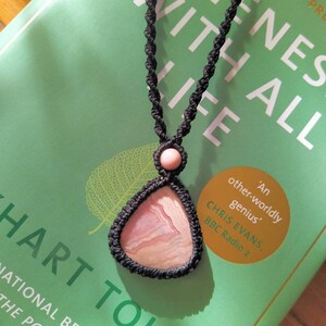 May include: A pink and white agate stone pendant on a black cord necklace. The pendant is in the shape of a triangle and has a natural, organic look. The necklace is lying on a green book with the text "ONENESS WITH ALL LIFE" and "HART TOLLE" visible.