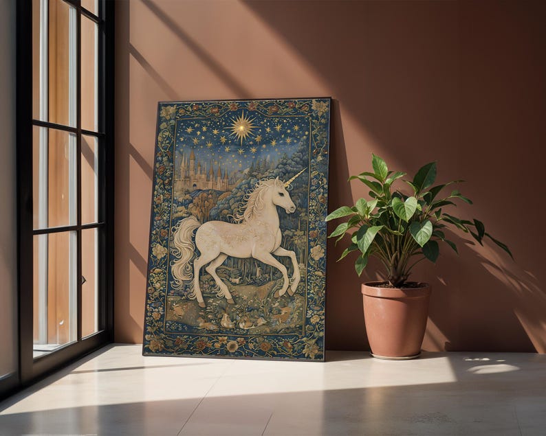 May include: A framed tapestry featuring a white unicorn with a golden horn, set against a night sky with stars and a cityscape. The tapestry has a blue background with floral borders and is displayed near a window and a potted plant.