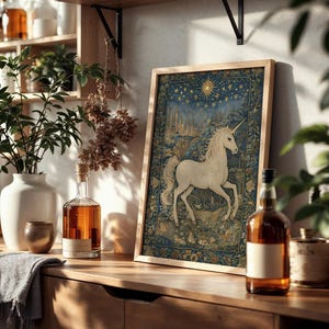 May include: A framed tapestry featuring a white unicorn against a blue and gold background. The artwork is displayed on a wooden surface, alongside a white vase with greenery, a bottle of amber liquid, and other decorative items.