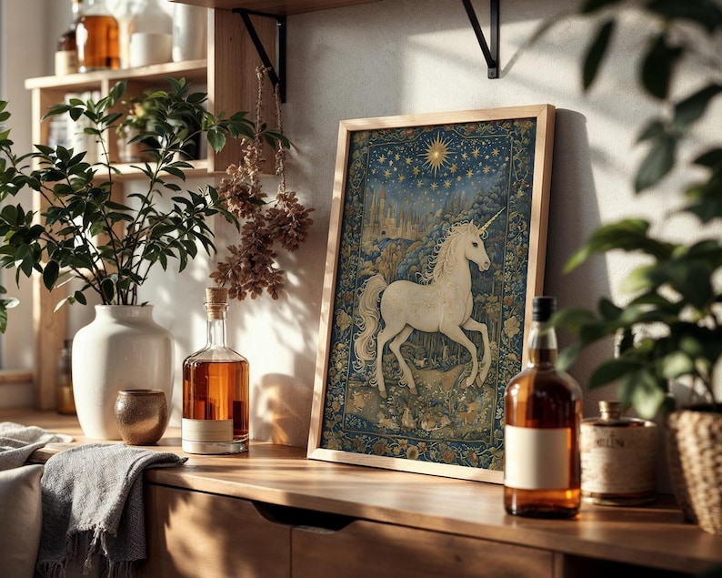 May include: A framed tapestry featuring a white unicorn against a blue and gold background. The artwork is displayed on a wooden surface, alongside a white vase with greenery, a bottle of amber liquid, and other decorative items.