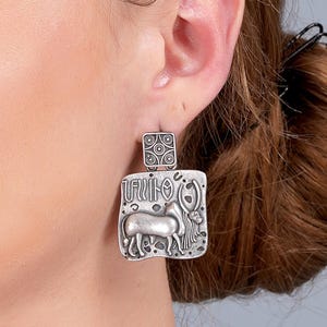 May include: Silver-toned rectangular earrings with a detailed relief design. The earrings feature a square top with a circular pattern and a larger rectangular bottom with a horse-like figure and other symbols. The earrings are worn on a person's ear.
