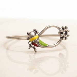May include: Silver bracelet with a colourful bird design. The bird has yellow, green, and red enamel accents. The bracelet has a loop design with a floral-shaped end and small dangling charms.