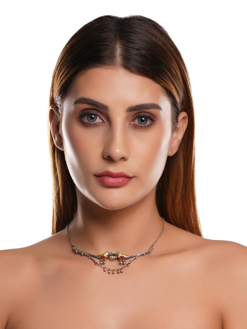 K&ouml;nnte beinhalten: Eine silberfarbene Halskette mit einem dekorativen Design mit roten und gelben Akzenten. Die Halskette hat eine zarte Kette und kleine, h&auml;ngende Elemente, die ein elegantes und detailliertes Schmuckst&uuml;ck schaffen.