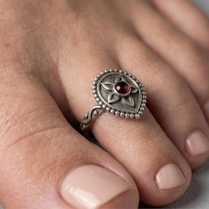 May include: A silver ring with a teardrop-shaped face featuring a central red gemstone and a floral design. The ring is adorned with small beaded details around the edge and has a decorative band.