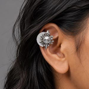 May include: A silver-toned ear cuff featuring a semi-circular flat plate design adorned with a central pyramid-shaped stud surrounded by a ring of small spherical beads and pointed accents, creating a textured, metallic, and geometric appearance.