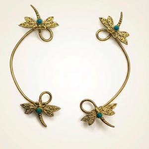 May include: Four gold-colored dragonfly ear cuffs. Each cuff features a dragonfly with intricate wing details and a small turquoise-colored stone. The dragonfly is attached to a curved wire that wraps around the ear.
