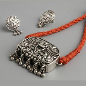 May include: A silver-toned pendant necklace featuring an ornate rectangular centerpiece with intricate embossed floral patterns and dangling spherical beads, suspended from a thick orange braided cord, accompanied by matching circular stud earrings.