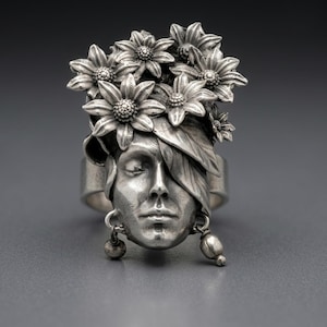 Puede incluir: Un anillo de plata con un rostro humano esculpido de ojos cerrados, adornado con una corona de flores y hojas detalladas. El rostro tiene pequeños dijes esféricos colgantes en las orejas, todo en un acabado de plata metálica pulida.