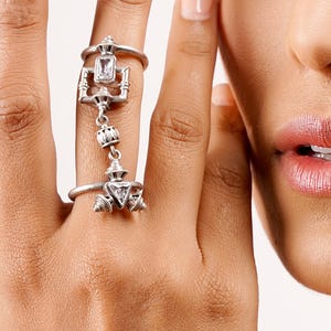 May include: A silver-toned ring set featuring three connected rings. One ring has a rectangular clear stone, another has a triangular clear stone, and the third is a simple band. The rings are connected by decorative chains.
