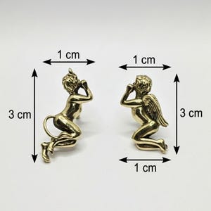 Può includere: Due piccole statuette color ottone, ciascuna di circa 1 cm di larghezza e 3 cm di altezza. Una raffigura un diavolo con corna e coda, l'altra un angelo con le ali. Entrambe sono inginocchiate con le mani vicino al viso.