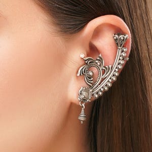 May include: A silver ear cuff with an ornate, floral design. The cuff curves around the ear, featuring a series of small, round beads and a dangling bell-shaped charm. The jewelry is worn on the ear, with the wearer's skin visible.