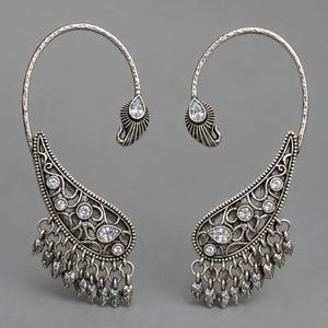 May include: A pair of silver-toned ear cuffs featuring an ornate paisley-shaped design with intricate filigree metalwork, clear faceted crystals, and a series of small dangling leaf-shaped charms along the bottom edge.